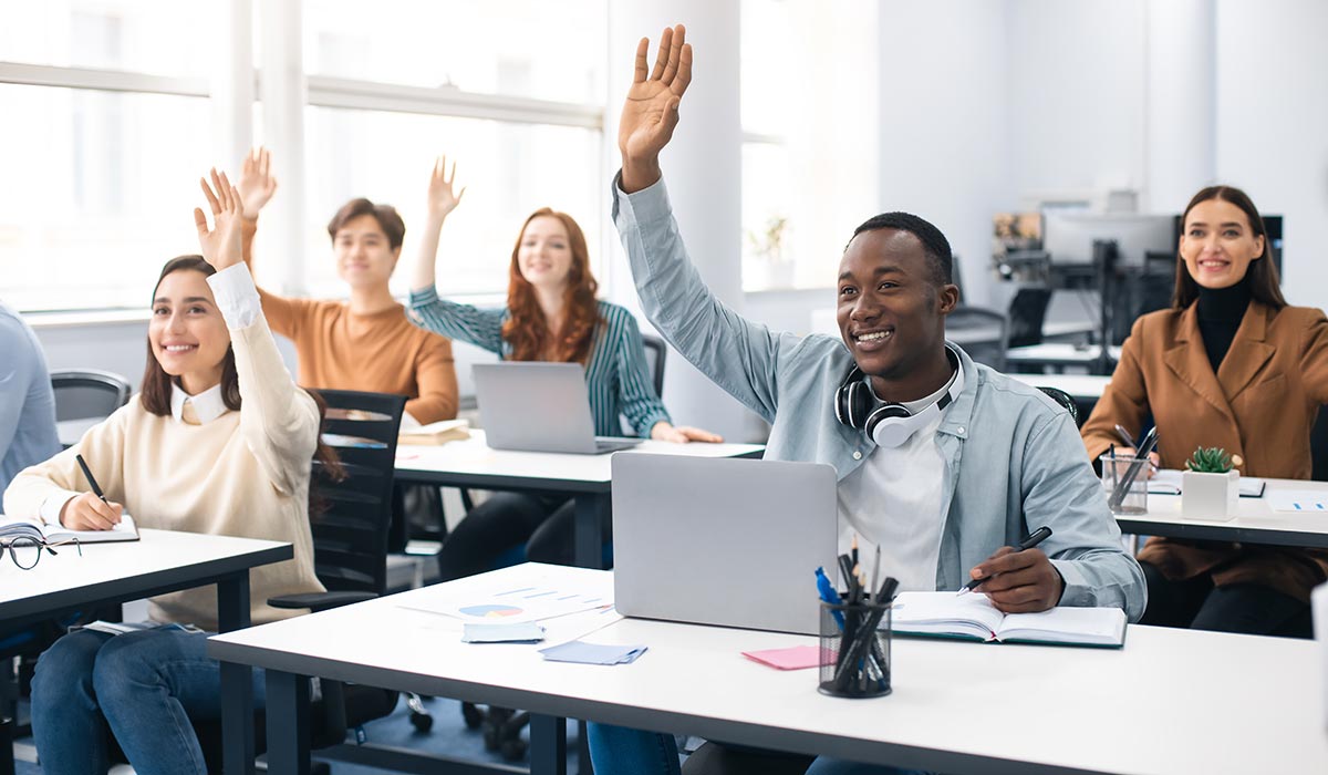 A group of adults in a training classroom raising their hands with the answer. Find out how an LMS can help with online and in-person training.